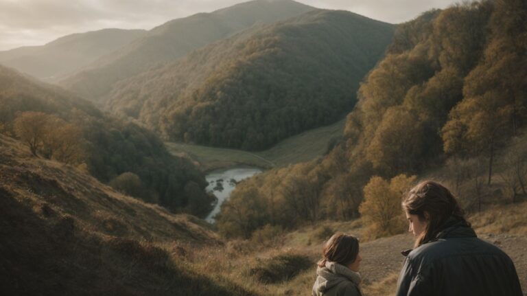 A peaceful outdoor scene with two individuals sitting on a hillside overlooking a river flowing through a lush valley surrounded by green and gold-colored trees during fall.